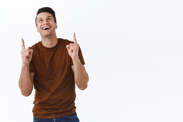 Portrait of cheerful happy caucasian masculine man looking and pointing fingers up showing funny advertisement found good deal enjoying view of something top standing white background