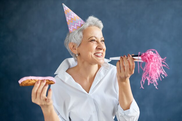 Portrait of cheerful happy middle aged woman in white shirt posing isolated with eclair and noisemaker in her hands, having fun at birthday party