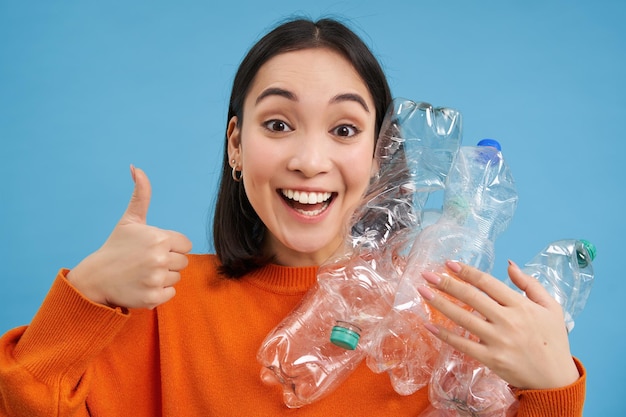 Portrait of cheerful korean girl shows thumbs up and plastic bottles recommends recycling at sorting
