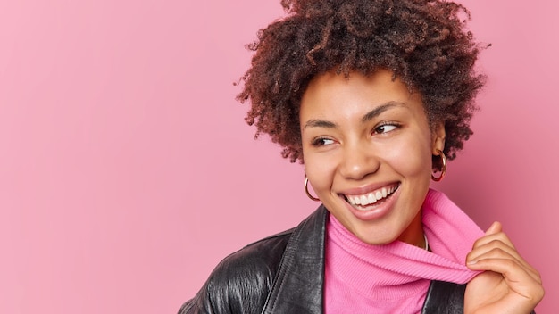 Portrait of cheerful lovely woman looks away with broad smile stretches collar of turtleneck wears leather jacket expresses positive emotions glad to notice something isolated over pink background
