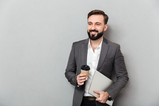 Portrait of cheerful male office worker posing on camera holding takeaway coffee and silver laptop, isolated over gray wall