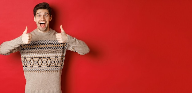Портрет веселого мужчины в рождественском свитере, похожего на что-то хорошее, показывает большой палец вверх и удивленно улыбается, хвалит новогоднюю вечеринку, стоя на красном фоне.