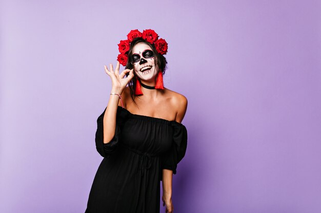 Portrait of cheerful Mexican with long earrings and red accessories in outfit for Halloween. Woman in great mood shows ok sign