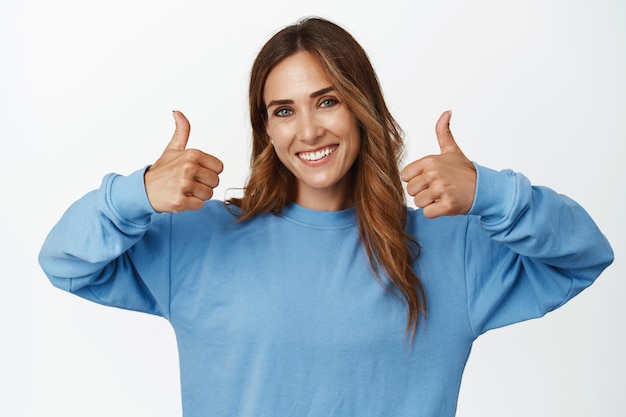 Portrait of cheerful middle aged 35 woman, showing thumbs up in approval, likes and agree, praise great work, nice job, recommending smth, standing against white background