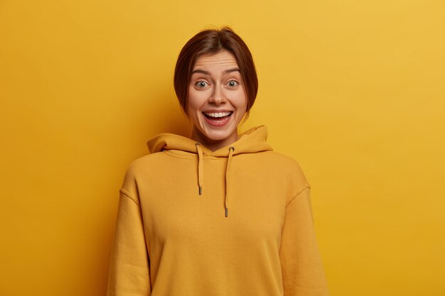 Portrait of cheerful millennial girl laughs happily, hears pleasant news, wears hoodie, has casual friendly conversation, beaming white smile, poses against yellow wall,