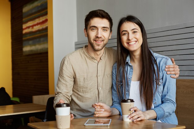 Portrait of cheerful perspective startupers pair with dark hair in casual clothes, sitting in cafe, smiling bright, drinking coffee. and hugging.