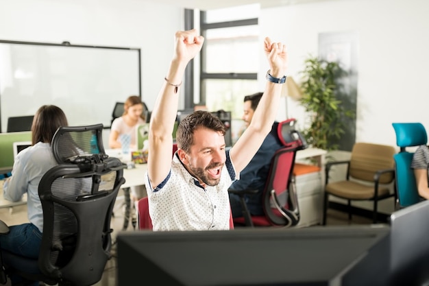 Portrait of cheerful positive man in causal outfit raising hands and celebrating achievement sitting in modern office with colleagues in background