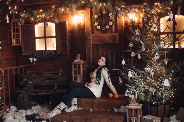 Портрет веселой и позитивной женщины с длинными темными волосами в свитере, джинсах и теплых носках, держащей завернутый подарок на Рождество, сидя под украшенной елкой и снегопадом