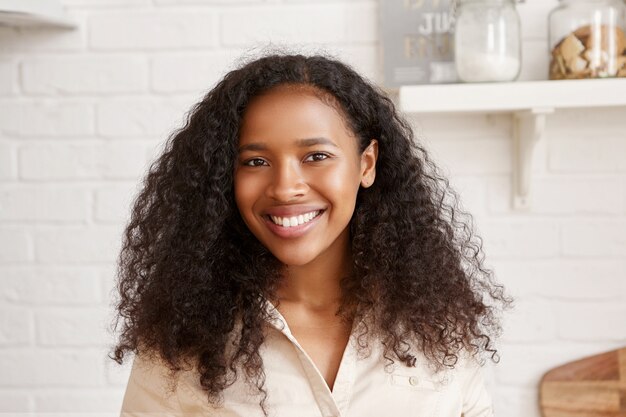 완벽한 하얀 치아, 방대한 검은 머리카락과 집에서 여가 시간을 보내는 반짝이는 검게 그을린 피부를 가진 쾌활한 긍정적 인 젊은 아프리카 여자의 초상화, 행복한 빛나는 미소로 부엌에서 포즈