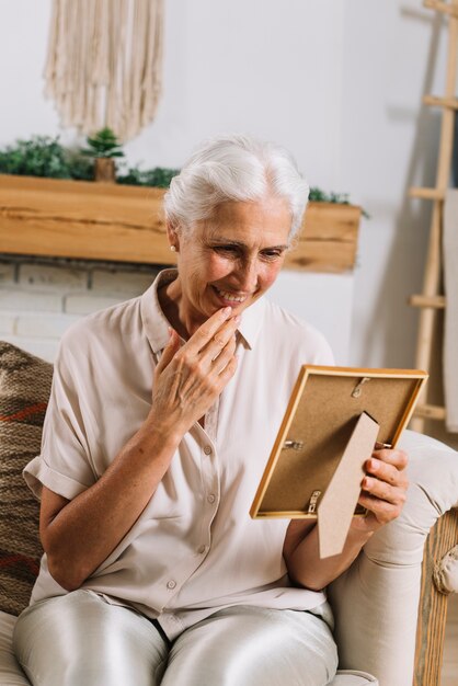 Portrait of cheerful senior woman sitting on sofa looking at photo frame