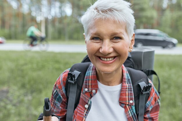 Portrait of cheerful short haired mature female hitchhiker carrying backpack and sleeping mat posing outdoors with high road and cars in background, going to spend vacations in wild nature