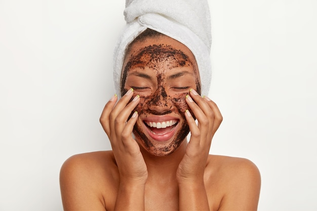 Free photo portrait of cheerful smiling dark skinned woman applies natural coffee mask, makes circular motions with hands and massages skin, stimulates facial blood supply, wears wrapped towel on head.