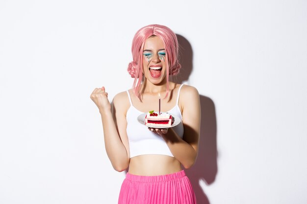 Portrait of cheerful smiling girl celebrating her birthday, wearing pink wig, holding b-day cake and shouting of joy, standing.