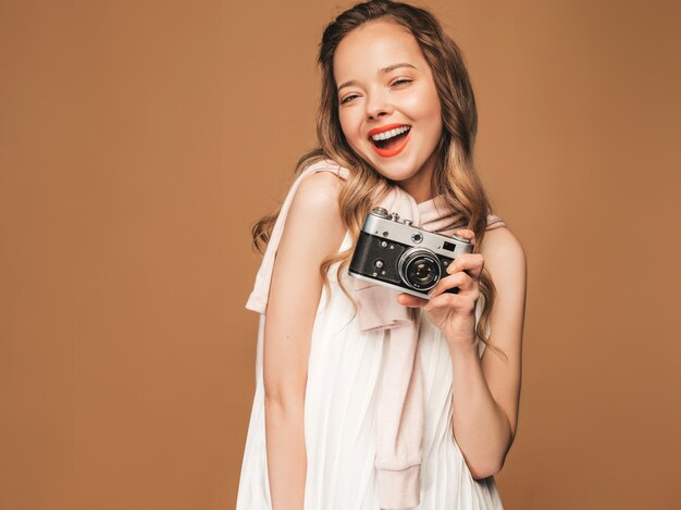 Portrait of cheerful smiling young woman taking photo  with inspiration and wearing white dress. Girl holding retro camera. Model posing