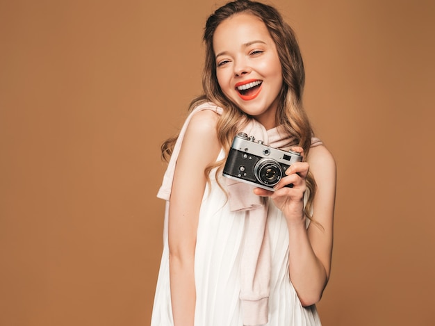 インスピレーションと写真を撮ると白いドレスを着て陽気な笑顔の若い女性の肖像画。レトロなカメラを保持している女の子。モデルのポーズ