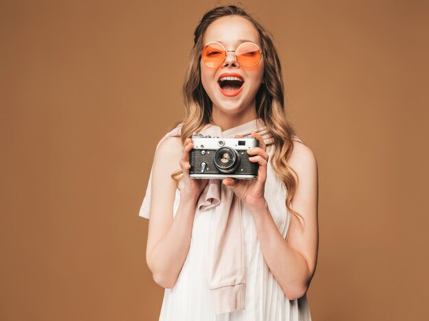 Portrait of cheerful smiling young woman taking photo  with inspiration and wearing white dress. Girl holding retro camera. Model in sunglasses posing