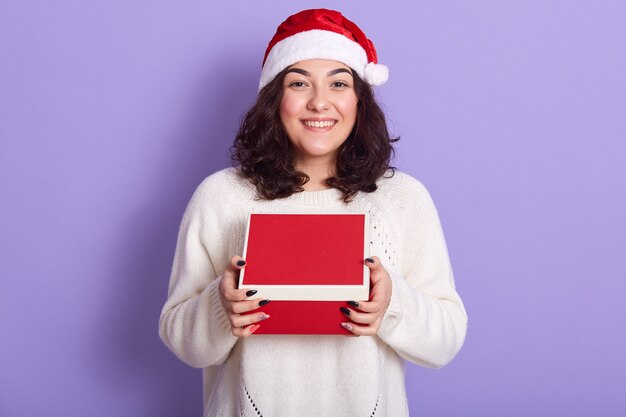 Портрет веселой нежной брюнетки, держащей коробку с подарком в обеих руках