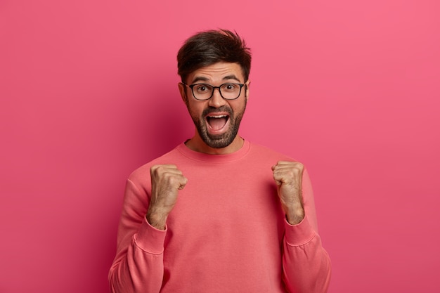 Ritratto di allegro uomo adulto con la barba lunga stringe i pugni con trionfo, si rallegra di un bel momento della vita, esclama positivamente, indossa occhiali trasparenti e maglione roseo, celebra la vittoria o il successo