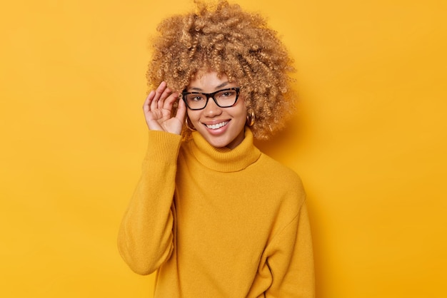 Portrait of cheerful woman keeps hand on rim of spectacles smiles joyfully wears casual jumper feels positive isolated over vivid yellow background stands pleased indoor. people and emotions