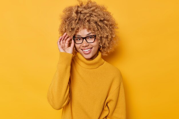 쾌활한 여성의 초상화는 안경 가장자리에 손을 대고 즐겁게 캐주얼 점퍼를 착용하고 선명한 노란색 배경 위에 긍정적으로 고립되어 실내를 기쁘게 생각합니다. 사람과 감정
