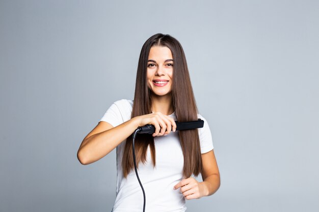 Portrait of cheerful woman using straightener for her curly hair preparing for event date holiday comfortable easy hairdo isolated on grey