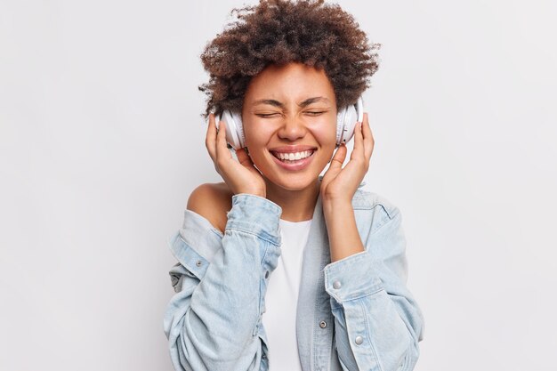 Portrait of cheerful woman with Afro hair keeps hands on stereo headphones keeps eyes closed smiles broadly shows white teeth enjoys pleasant music isolated over white wall