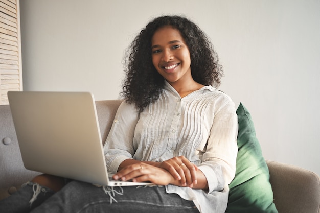 Free photo portrait of cheerful young african woman in jeans and shirt smiling broadly while surfing internet on generic portable computer, enjoying high speed wireless connection in living room