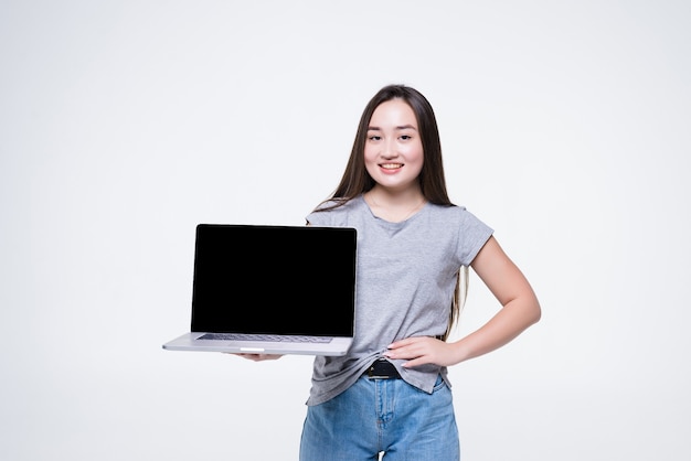 白い壁の上に孤立して座っている間空白の画面のラップトップコンピューターに指を指している陽気な若いアジアの女性の肖像画