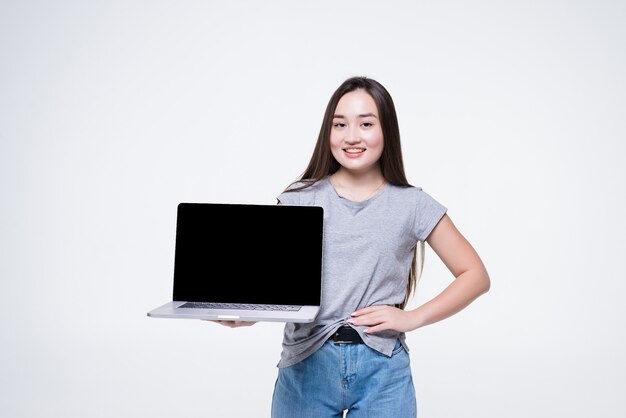 Portrait of a cheerful young asian woman pointing finger at blank screen laptop computer while sitting isolated over white wall