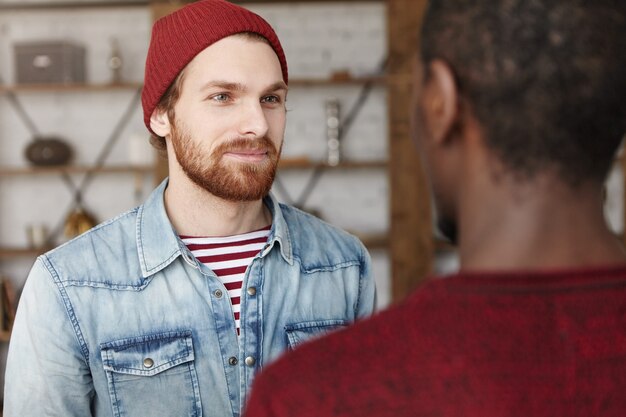 Portrait of cheerful young bearded Caucasian hipster wearing trendy hat and denim jacket