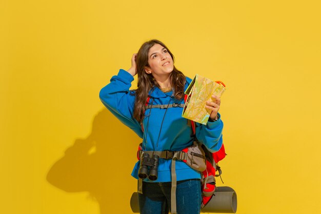Portrait of a cheerful young caucasian tourist girl with bag and binoculars isolated on yellow studio background.