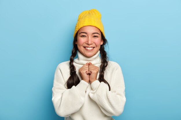쾌활한 어린 소녀의 초상화는 손을 함께 유지하고, 카메라에 즐겁게 미소를 짓고, 긍정적 인 소식을 즐기고, 노란 모자와 흰색 대형 스웨터를 입습니다.