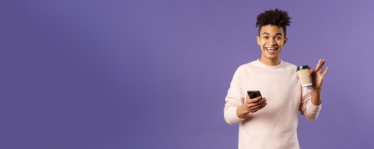 Free Photo portrait of cheerful young male student with cup of coffee and mobile phone waving hand to say hi sm