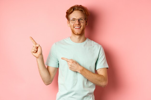 Portrait of cheerful young man with red hair wearing glasses pointing fingers at upper left corner a...
