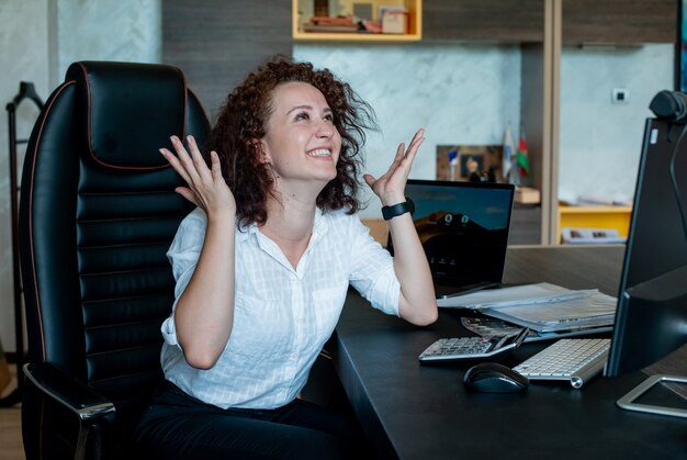 Портрет веселой молодой женщины офисного работника, сидящей за офисным столом, счастливой и взволнованной с поднятыми руками, весело улыбаясь в офисе