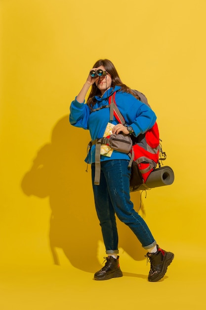 黄色のスタジオの壁に分離されたバッグと双眼鏡で陽気な若い観光の女の子の肖像画