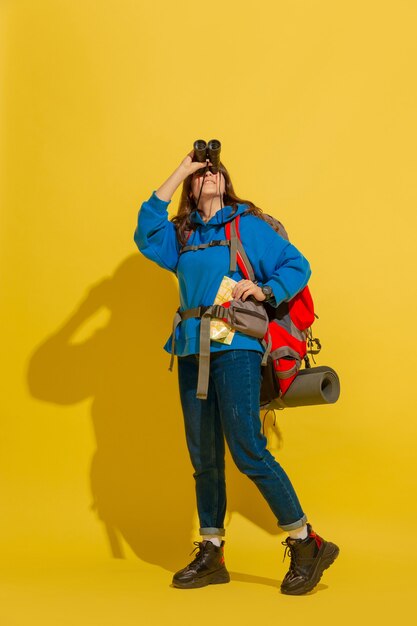 Portrait of a cheerful young tourist girl with bag and binoculars isolated on yellow studio wall