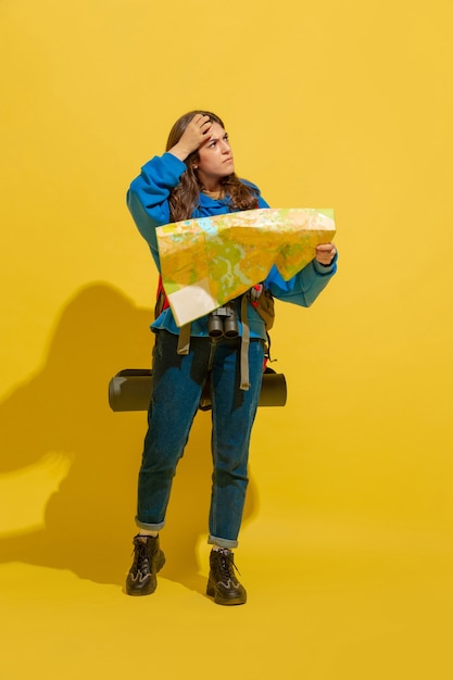 Free photo portrait of a cheerful young tourist girl with bag and binoculars isolated on yellow studio wall
