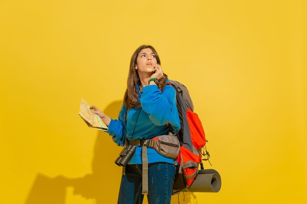 Portrait of a cheerful young tourist girl with bag and binoculars isolated on yellow studio wall