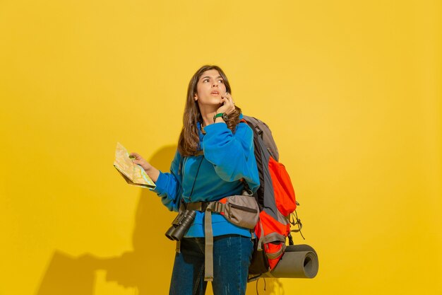 Портрет веселой молодой туристической девушки с сумкой и биноклем, изолированной на желтой стене студии