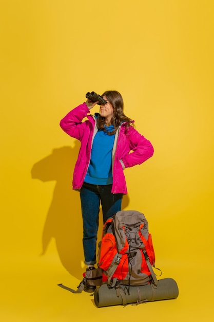 Portrait of a cheerful young tourist girl with bag and binoculars isolated on yellow studio wall