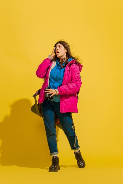 Portrait of a cheerful young tourist girl with bag and binoculars isolated on yellow studio wall