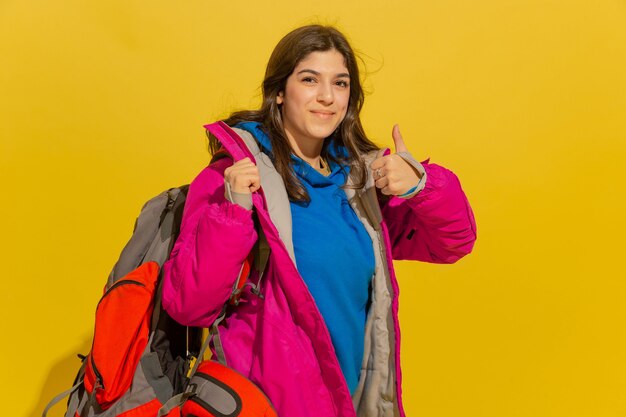 Portrait of a cheerful young tourist girl with bag and binoculars isolated on yellow studio wall