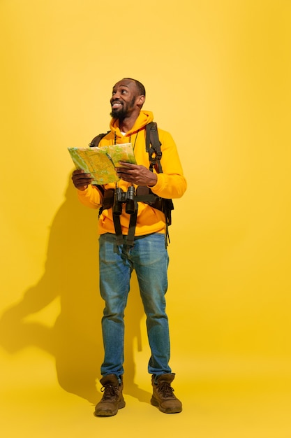 Portrait of a cheerful young tourist guy with bag and binoculars isolated on yellow studio wall