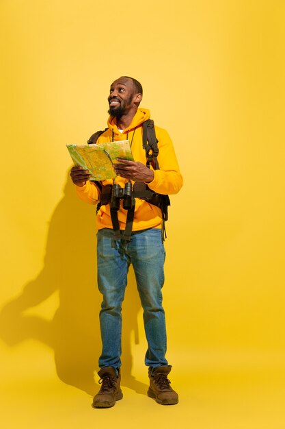 Portrait of a cheerful young tourist guy with bag and binoculars isolated on yellow studio wall