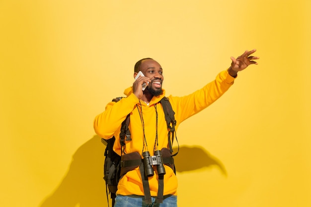 Portrait of a cheerful young tourist guy with bag and binoculars isolated on yellow studio wall