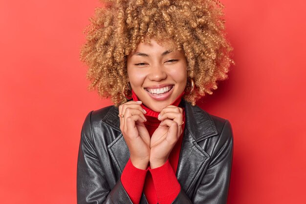 Portrait of cheerful young woman keeps hands on collar of jumper smiles broadly shows white perfect teeth dressed in leather jacket isolated over red background. Authentic human emotions concept