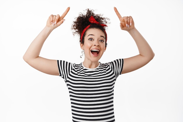 Portrait of cheerful young woman laughing, pointing fingers up, showing promotional text on top, advertising something, standing against white background.