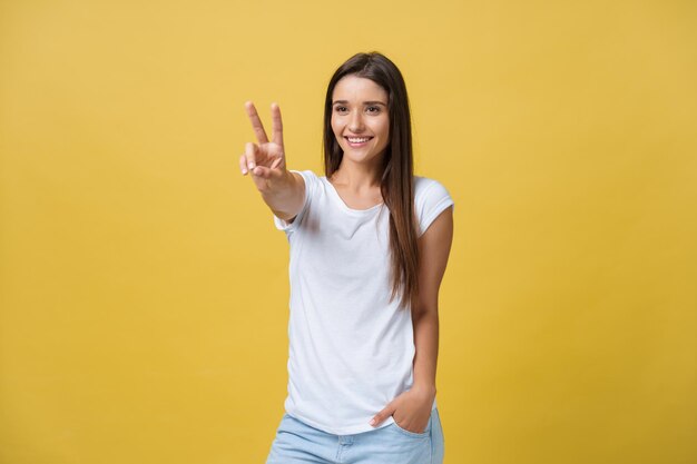 黄色の背景の上に2本の指または勝利のジェスチャーを示す陽気な若い女性の肖像画