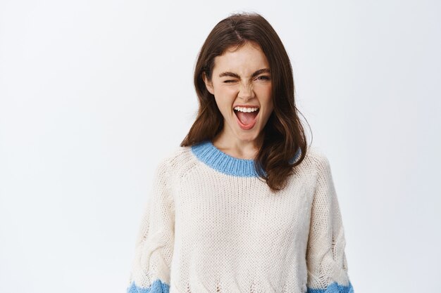 Портрет веселой молодой женщины в свитере, показывающей язык и подмигивающей возбужденно, наслаждающейся вечеринкой, выражающей веселье и положительные эмоции, стоя у белой стены